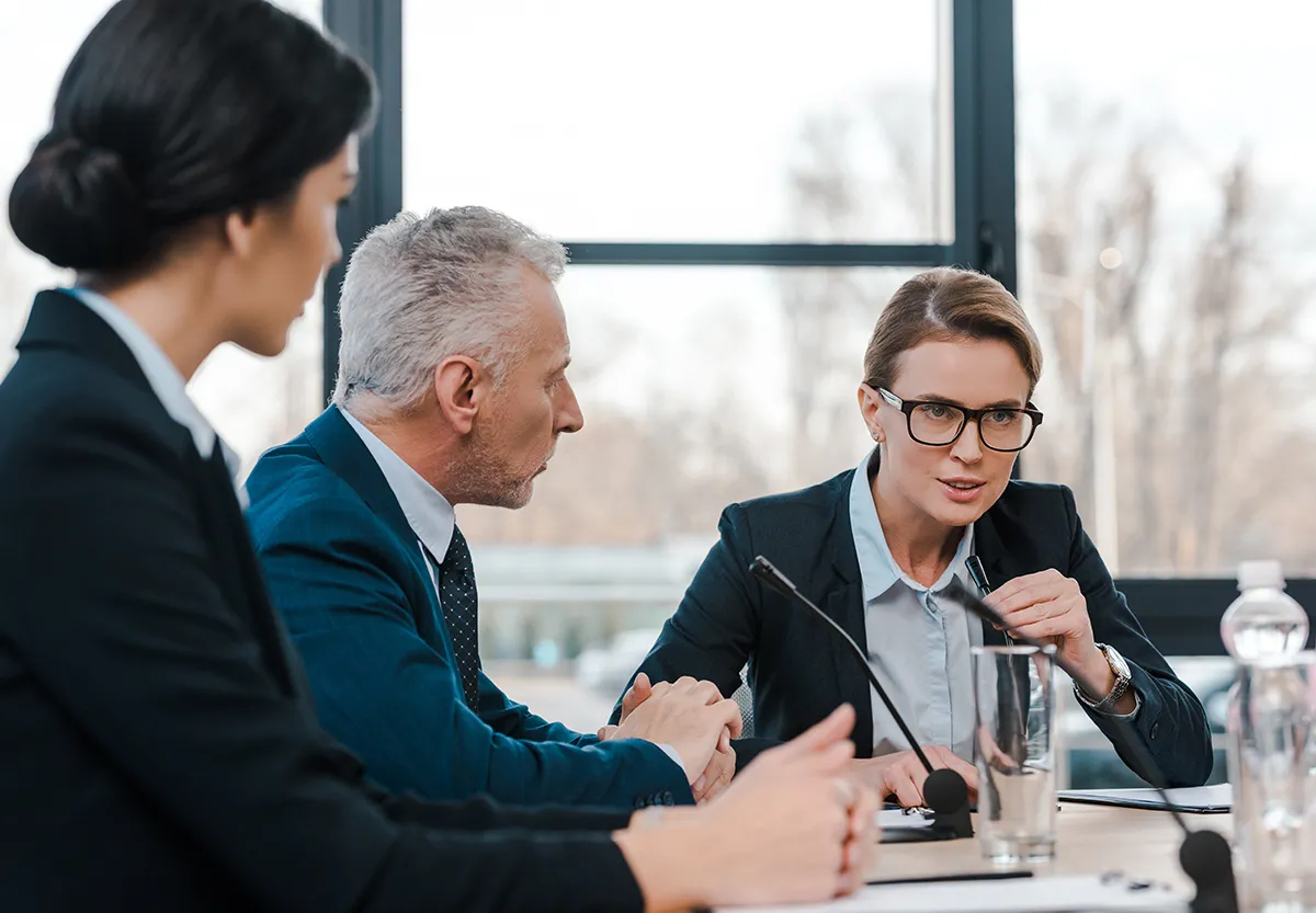 Three people in formal attire in a focused conversation around a conference table.