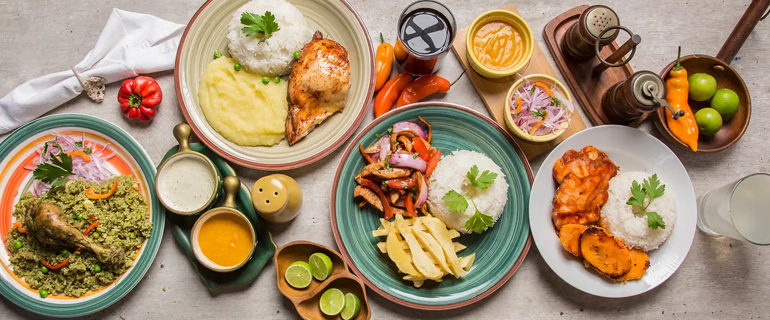 Overhead flat-lay of Peruvian dishes: arroz con pato, lomo saltado with chips, grilled fish, ají amarillo sauces, red onion salad, and fresh limes.